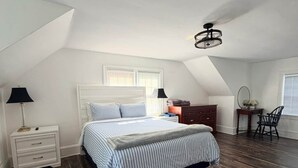 A bedroom with a sloped ceiling, a bed with striped bedding, a nightstand, a dresser, a chair, and a desk with a lamp. - Magnolia Breeze (Norfolk)