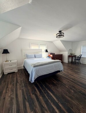 Un dormitorio con techo inclinado, una cama con ropa de cama a rayas, una mesita de noche, un aparador, una silla y un escritorio con una lámpara.