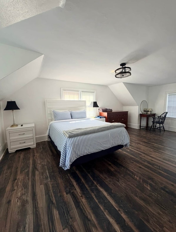 A bedroom with a sloped ceiling, a bed with striped bedding, a nightstand, a dresser, a chair, and a desk with a lamp.