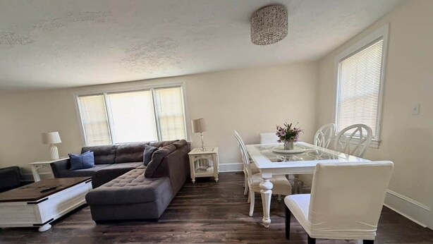 A living room with a sectional sofa, a dining table with chairs, and a small side table with a lamp. - Magnolia Breeze (Norfolk)