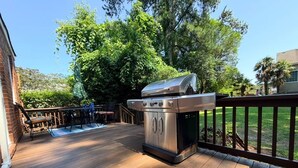 An outdoor deck with a stainless steel grill, wooden deck flooring, and a fenced area with a view of a building and trees. - Magnolia Breeze (Norfolk)