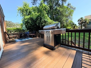 Una terraza al aire libre con una parrilla de acero inoxidable, suelo de madera y una zona vallada con vistas a un edificio y árboles.