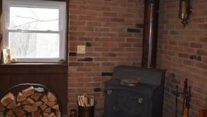 A room with a brick wall, a window, a fireplace with a chimney, a log stack, and a guitar leaning against the wall.