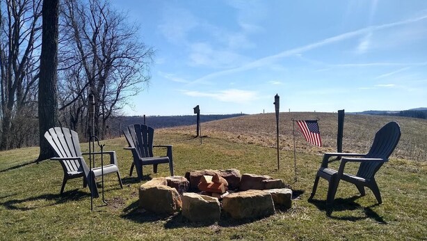 A fire pit with four Adirondack chairs around it, set on a grassy field with trees and a flag in the background.