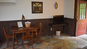A room with a wooden table, chairs, a television, a wall-mounted air conditioner, a decorative wall piece, and a red door.