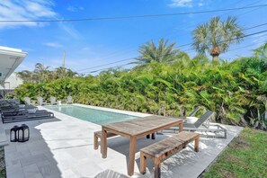 A poolside area with a wooden table and benches, surrounded by lush greenery and a clear blue sky.