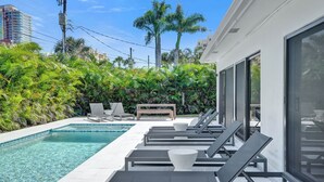 Un área de piscina moderna con sillones reclinables y un banco, rodeada de exuberante vegetación y palmeras.