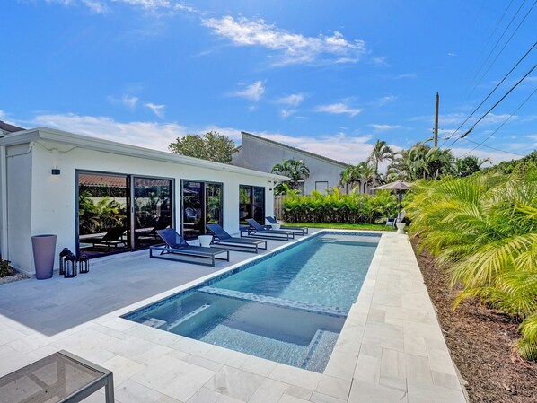 A modern pool area with a clear blue pool, lounge chairs, and a patio with a view of the house.