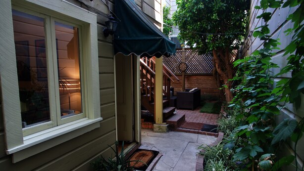 A narrow outdoor pathway leading to a wooden door with a green awning, surrounded by potted plants and a wooden fence. - Lovely 1 Bedroom Garden flat in Victorian Building (San Francisco)