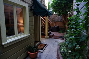 A narrow outdoor pathway leading to a wooden door with a green awning, surrounded by potted plants and a wooden fence.