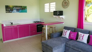 Une cuisine de taille réduite avec des meubles roses, une cuisinière à gaz et un four à micro-ondes. On y trouve un canapé gris avec des coussins et un petit tabouret. Un tableau représentant une cascade est accroché au mur, et une horloge est visible.