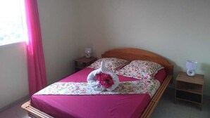 Un lit bien fait, avec une couvre-lit rose et des coussins à motifs floraux, une tête de lit en bois, une table de chevet avec une lampe et un appareil de climatisation fixé au mur.