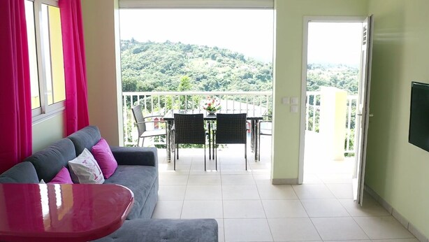 Une pièce avec un canapé gris, une table rouge et un balcon équipé d’une table et de chaises.