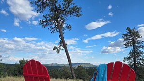 Dos sillas Adirondack rojas, una con una toalla azul, sobre un suelo cubierto de astillas de madera, con un cielo azul despejado y un árbol al fondo.
