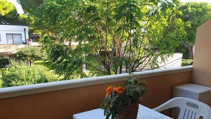 Ein Balkon mit einem weißen Tisch und Stühlen, einem Topf mit orangefarbenen Blüten sowie Blick auf Grünflächen und Gebäude.
