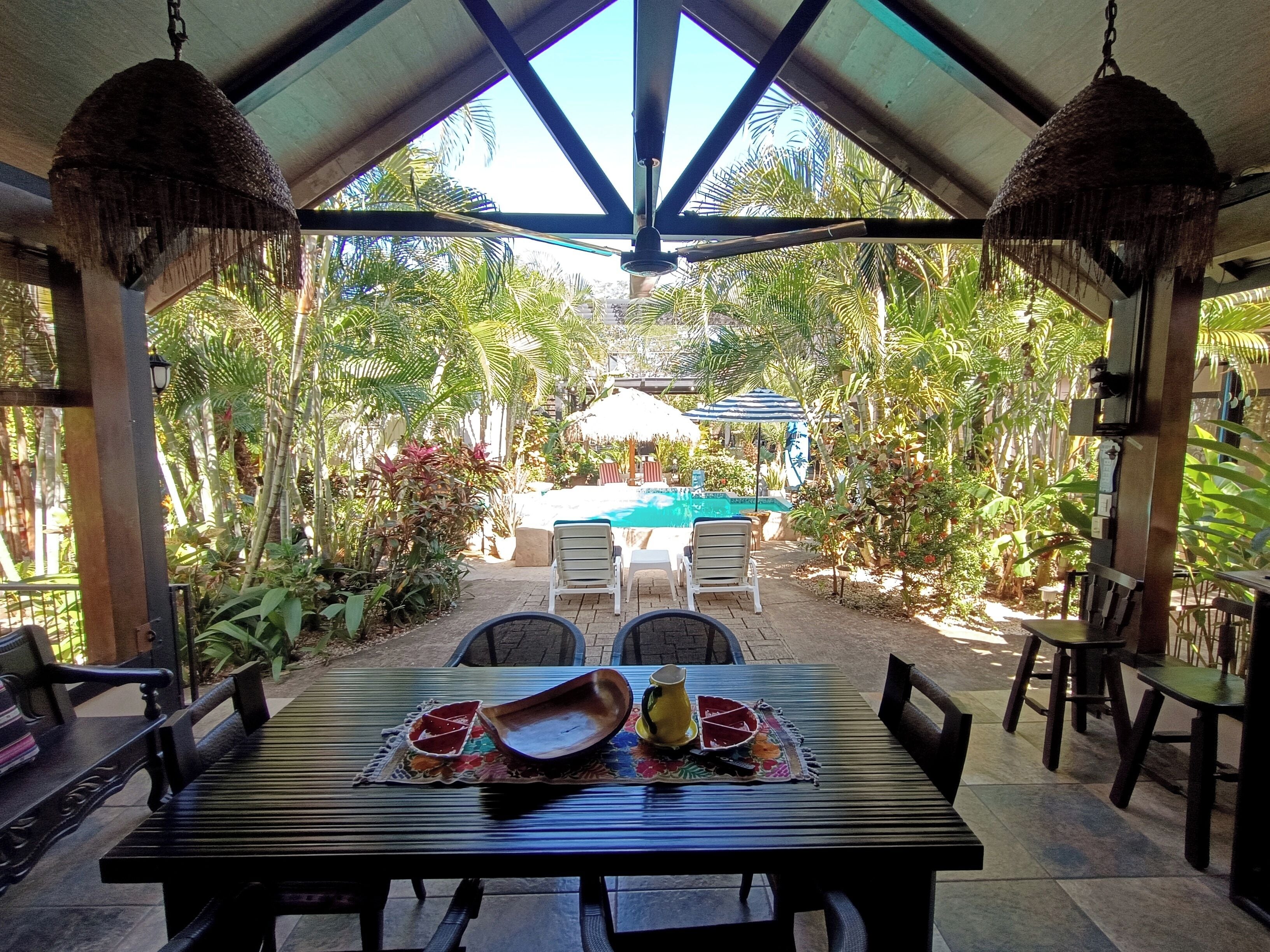 An outdoor patio with a pool, surrounded by lush greenery and a covered seating area.