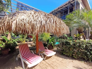 Relax in a tranquil outdoor space with two lounge chairs, a thatched umbrella, and lush greenery.