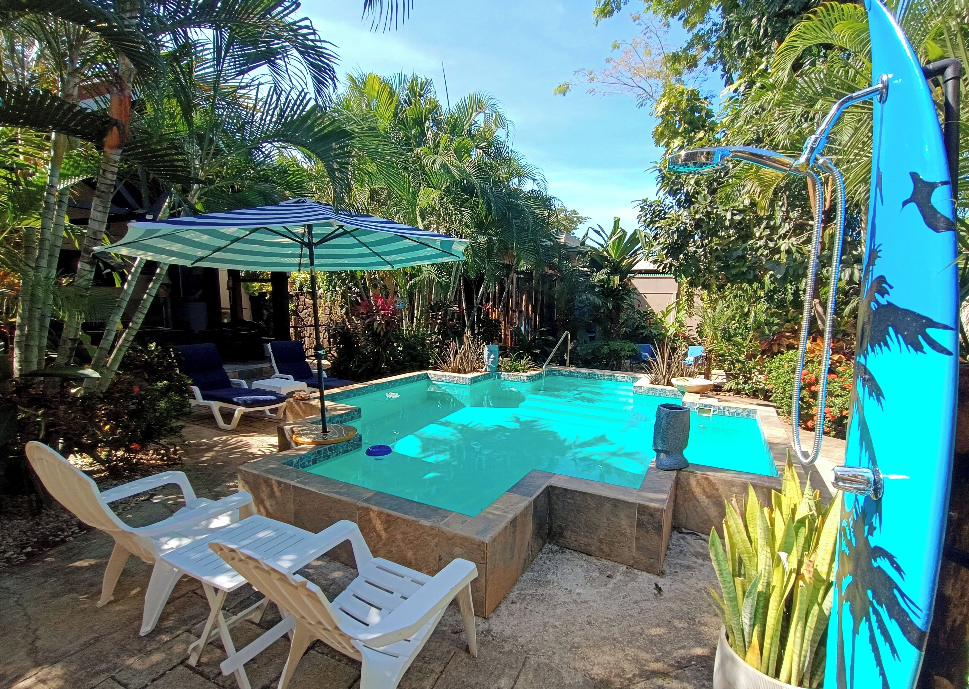 A swimming pool with lounge chairs and a covered area, surrounded by tropical plants.
