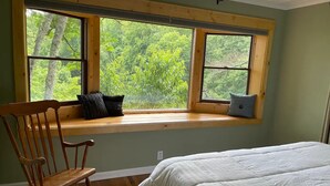 Un dormitorio con un ventanal grande, una mecedora de madera y una cama con ropa blanca.