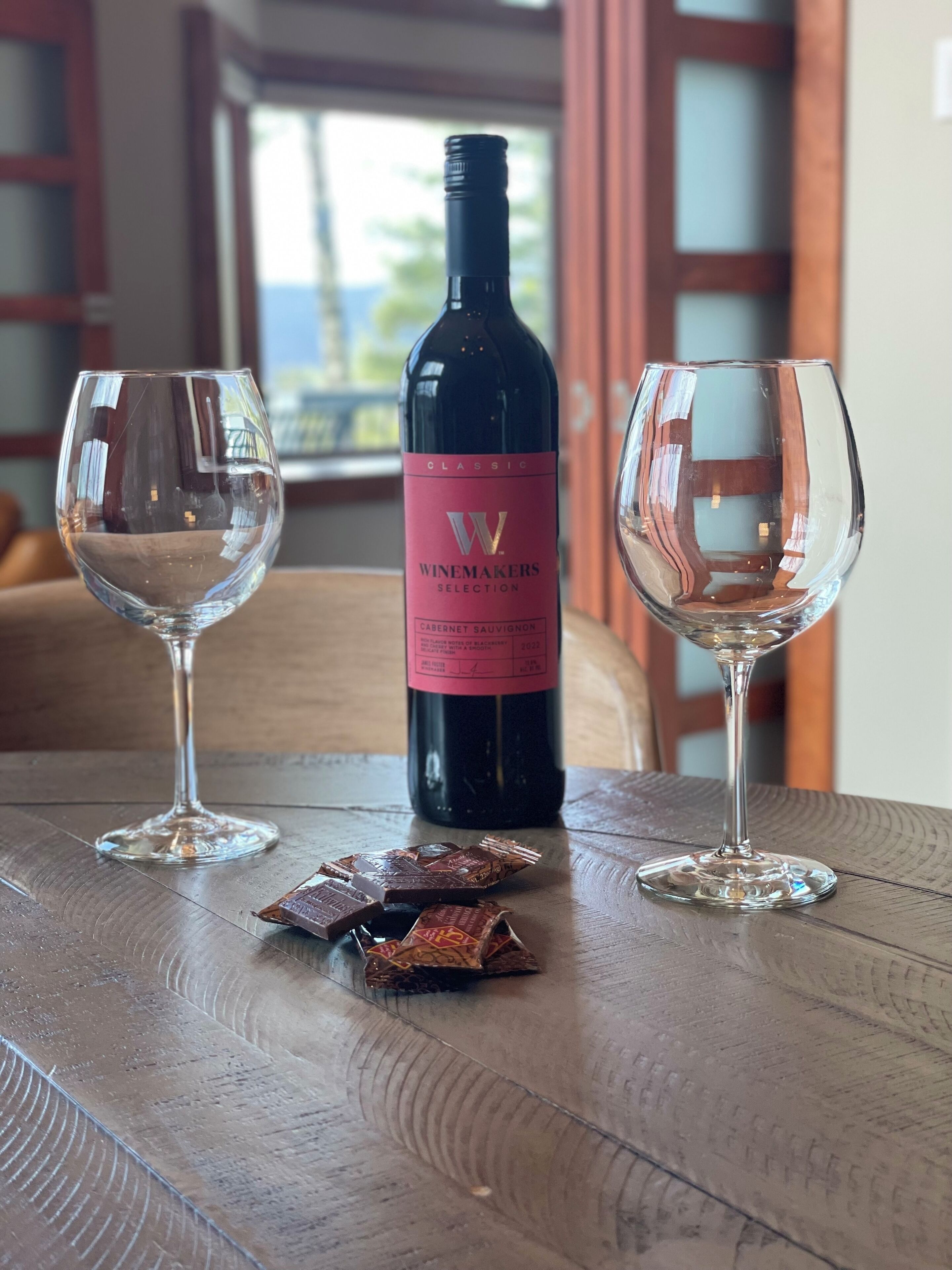 Uma garrafa de vinho com etiqueta rosa, dois copos de vinho vazios e uma barra de chocolate sobre uma mesa de madeira.