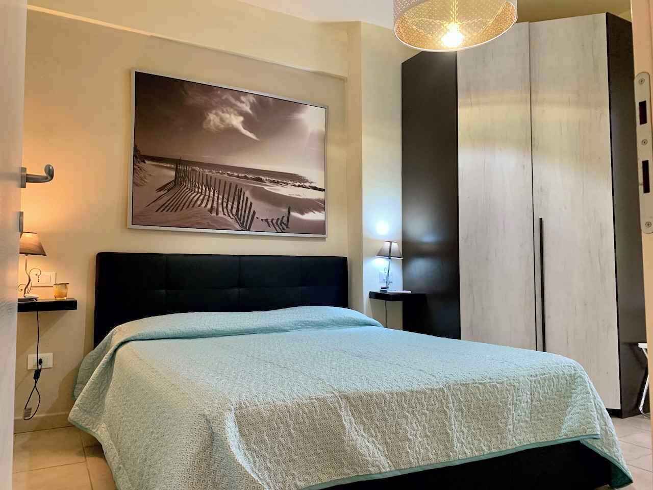 A bedroom with a bed, a nightstand, a lamp, a wardrobe, and a painting on the wall.