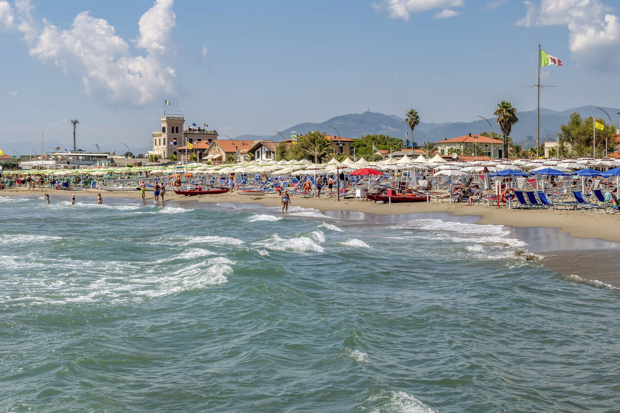 A beach with umbrellas and lounge chairs, a building with a flag, and mountains in the background.