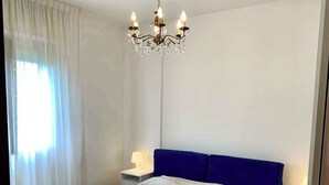 A bedroom with a large bed, a chandelier, a nightstand with a lamp, and a window with curtains. - Holiday apartment with air conditioning (Massa)