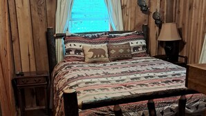 Ein Zimmer mit Holzboden, einem Bett, einem Nachttisch, einer Lampe und einem Fenster mit Vorhängen.