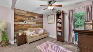Ein gemütliches Wohnzimmer mit einer Wand in Holzoptik, einem Deckenventilator, einem Bücherregal, einem Sofa mit gemusterten Kissen und einem Fenster mit Vorhängen.