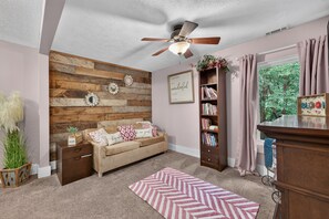 A cozy living room with a wooden accent wall, a ceiling fan, a bookshelf, a sofa with patterned cushions, and a window with curtains. - Rare find! Spacious home near wedding venues & CB/OMA. Perfect for entertaining! (Glenwood)
