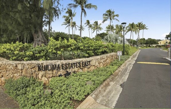 A stone wall with the text "KUIHLIMA ESTATES EAST" and a street with palm trees and a clear sky. - Turtle Bay Condo Lilinoe (Kahuku)