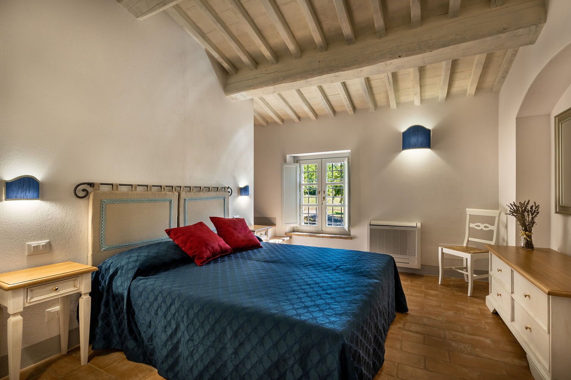 Ein Schlafzimmer mit einem Bett, einem Nachttisch, einer Kommode, einem Fenster und einer Wandlampe.