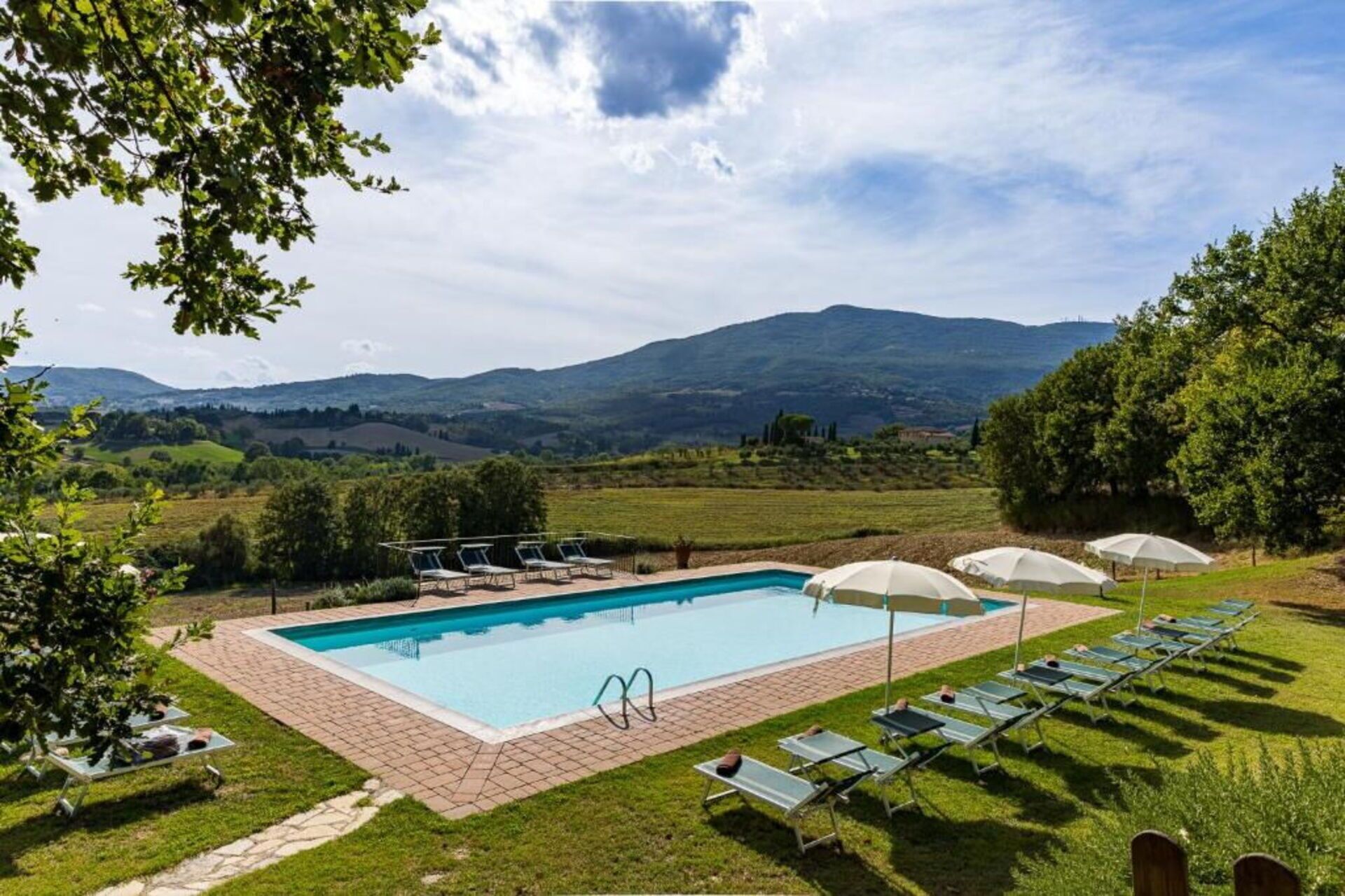Ein rechteckiger Pool mit Backsteinrand, umgeben von Gras und Bäumen. Liegestühle und Sonnenschirme befinden sich rund um den Pool.