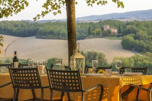 Une table à manger en plein air, avec des chaises, une bouteille de vin et des verres, le tout disposé devant un paysage pittoresque d’une vallée et de bâtiments éloignés.