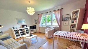 A living room with a sofa, a coffee table, a dining table with a checkered tablecloth, a bookshelf, a television, and a window with curtains. - Hook - Holiday apartment Hook - with south-facing terrace, very quiet location (Midlum)