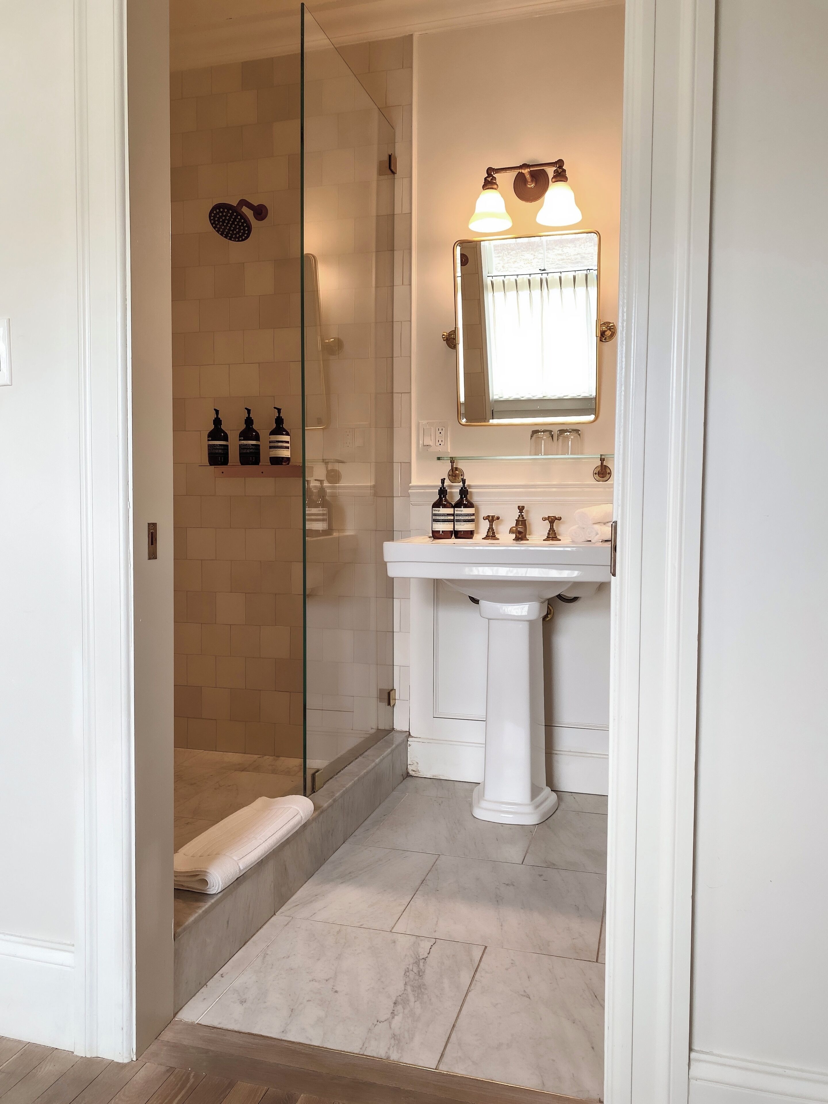 A bathroom with a glass shower enclosure, a pedestal sink, and a mirror with lights.