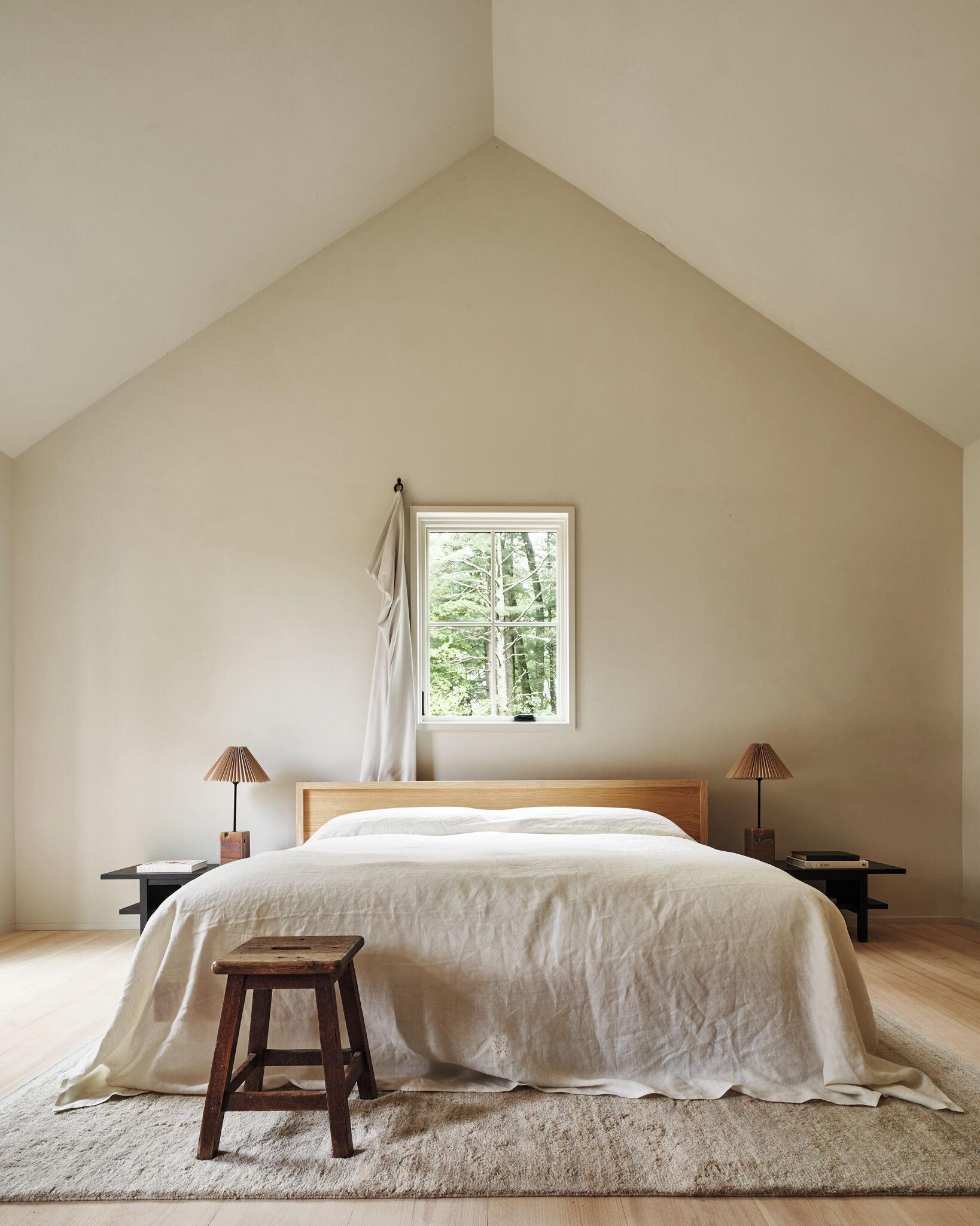 A bedroom with a sloped ceiling, a large window with a view of trees, a bed with white bedding, two bedside tables with lamps, and a small stool.