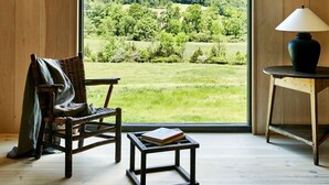 Una habitación con paredes y techo de madera, un ventanal con vistas a un paisaje verde, una silla de mimbre, una mesita con un libro y una lámpara sobre un taburete de madera.