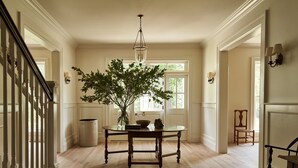 Una habitación amplia con piso de madera, una mesa redonda con un florero de follaje verde y una lámpara colgante.