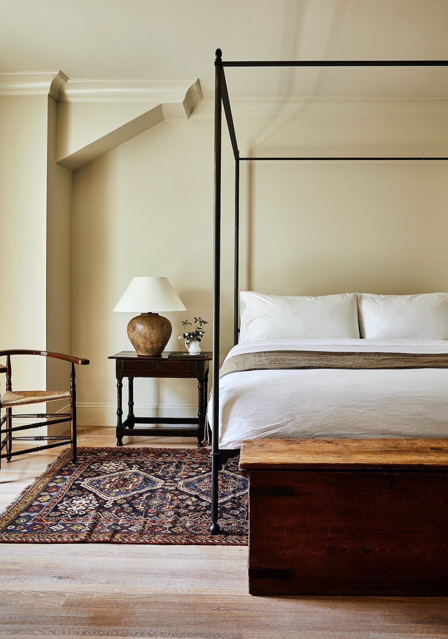 Un dormitorio con una cama con dosel, una mesita de noche de madera con lámpara, una silla y una alfombra estampada.