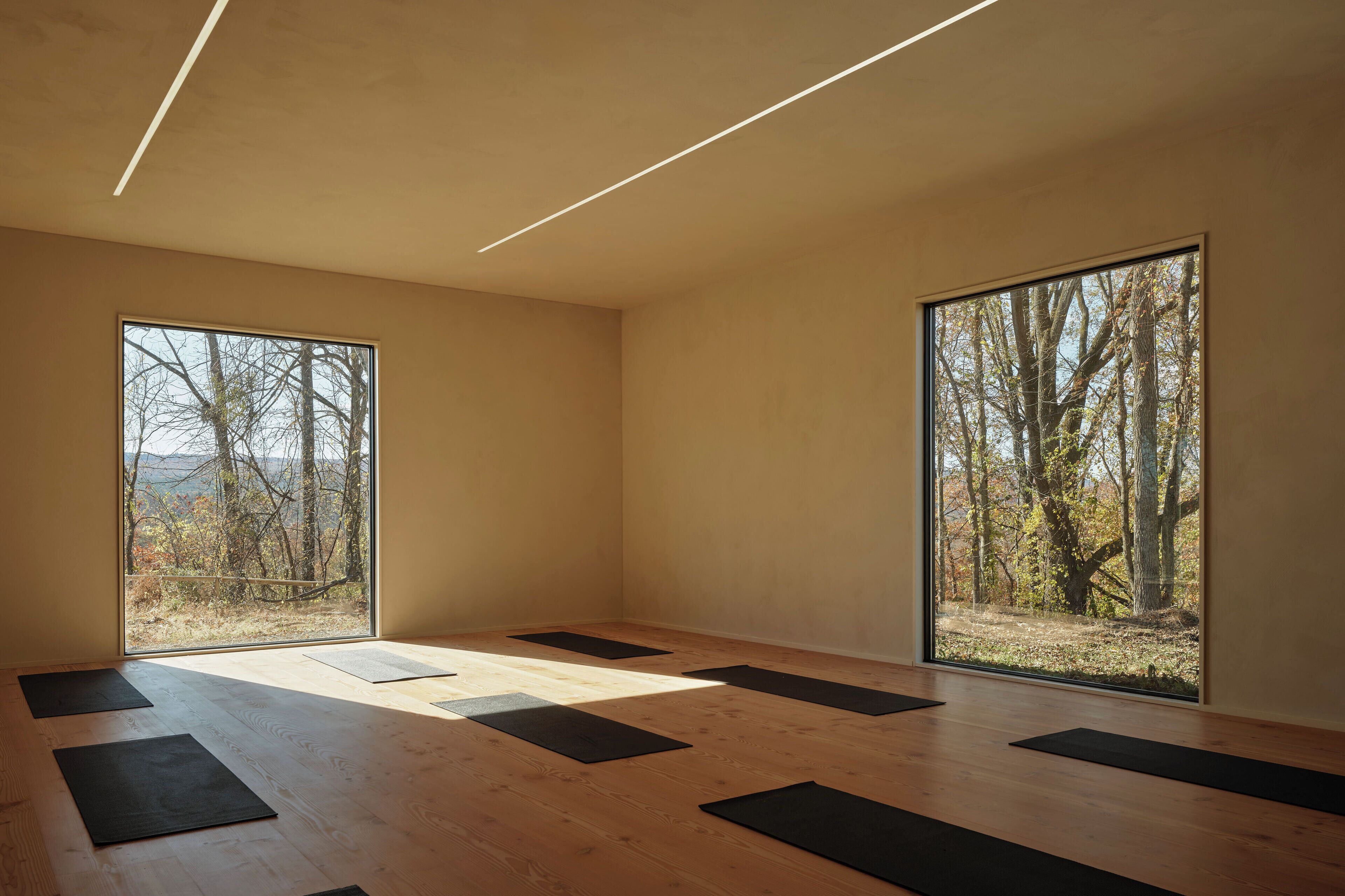 A spacious room with large windows, wooden flooring, and black mats arranged for yoga or stretching.