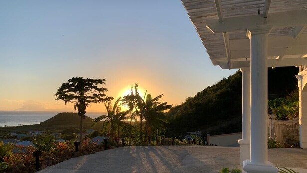 Un camino pavimentado conduce a un edificio con un porche cubierto, con vistas a un paisaje costero con palmeras y una montaña al fondo, bajo un atardecer.