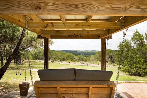 A wooden swing bench under a covered area with a view of a grassy landscape and trees.