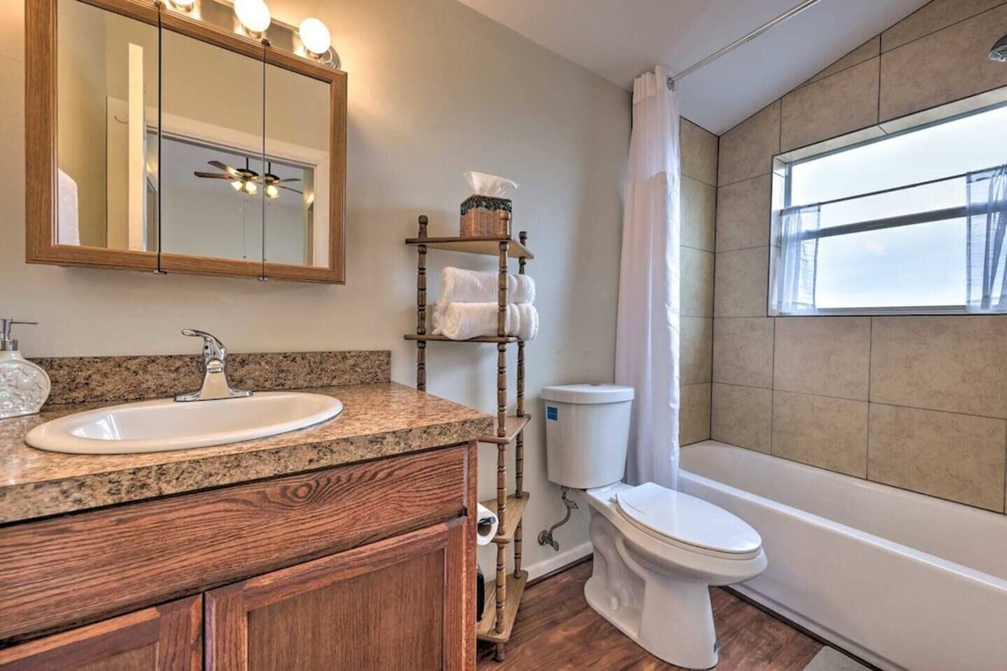 A bathroom with a granite countertop sink, wooden vanity, a mirror with lights, a toilet, and a window with a view.