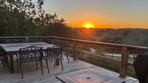 A wooden deck with a table and chairs, overlooking a sunset. - Las Estrellas Lots of privacy with stunning views, 2 decks, private hot tub! (Wimberley)
