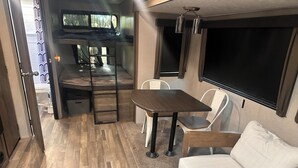 Een compact RV-interieur met een kitchenette, een zithoek met tafel en stoelen, een bank en een groot raam.