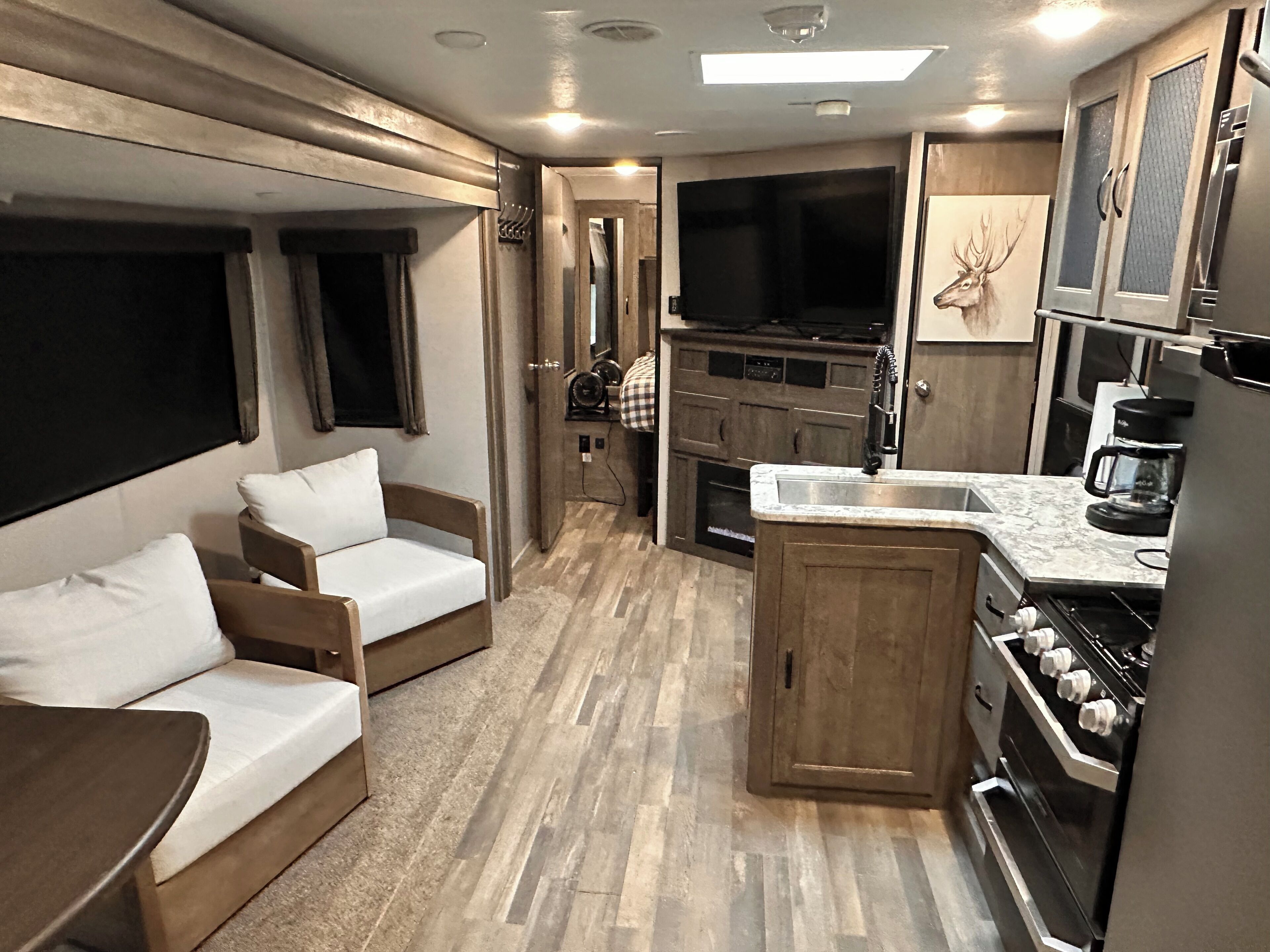 Interior d'una caravana compacta amb cuineta, zona d'estar amb sofà i taula, i un televisor gran fixat a la paret.