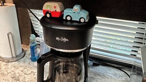 Een koffiezetapparaat met een autovormig deksel, een witte waterkoker en een blauwe fles op een keukenblad.