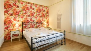 Et soveværelse med blomstret tapet, en seng med metalramme, et natbord med lampe og et vindue med gardiner.