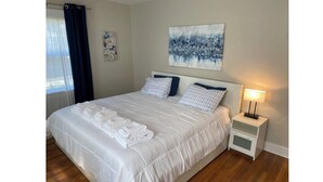 Een net opgemaakt bed met witte lakens en een opgevouwen handdoek, een nachtkastje met een lamp, en een schilderij aan de muur.